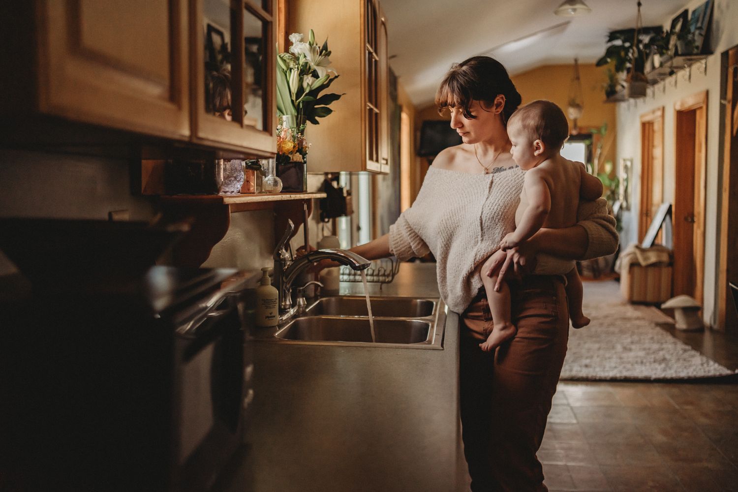 Parent multitasking in kitchen while holding baby in warm ambient lighting.