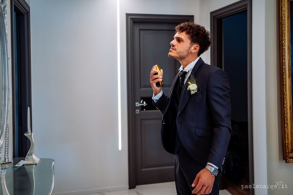 Groom in classic navy suit adjusts tie while standing in elegant interior hallway.