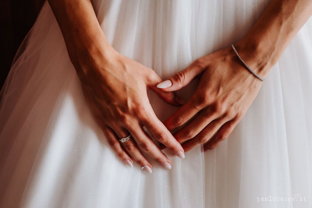 Close-up of hands resting on white wedding dress fabric with delicate jewelry details.