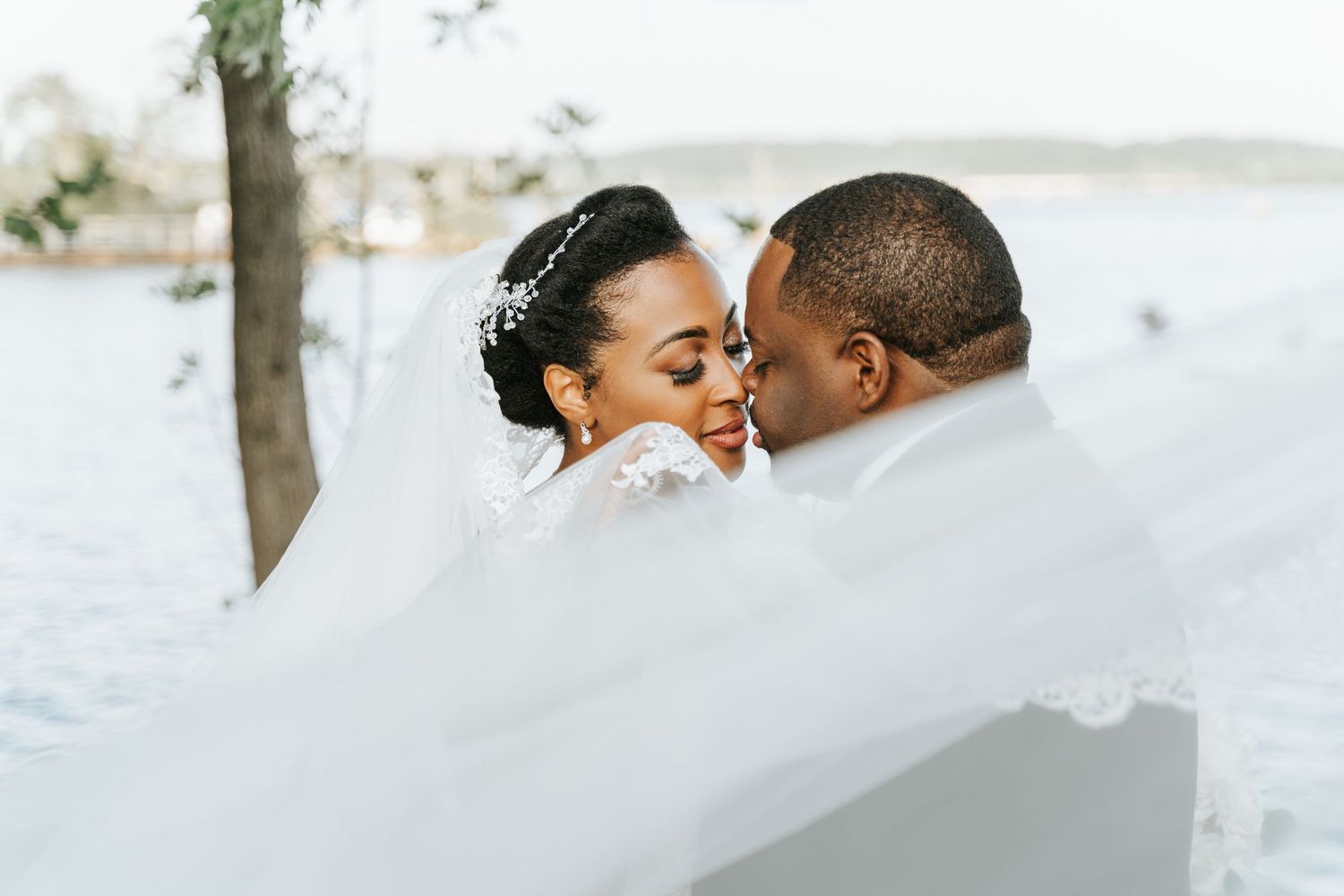 Wedding couple sharing an intimate moment under flowing white veil by the waterfront.