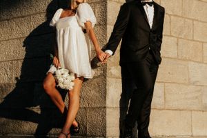 Couple holding hands against stone wall with dramatic shadows and formal attire.