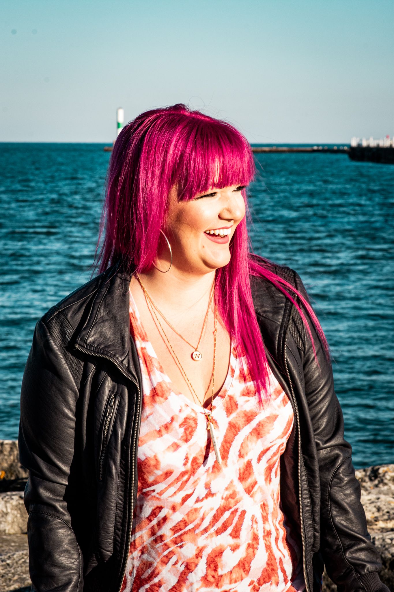 Woman with vibrant pink hair laughing by the ocean wearing leather jacket.