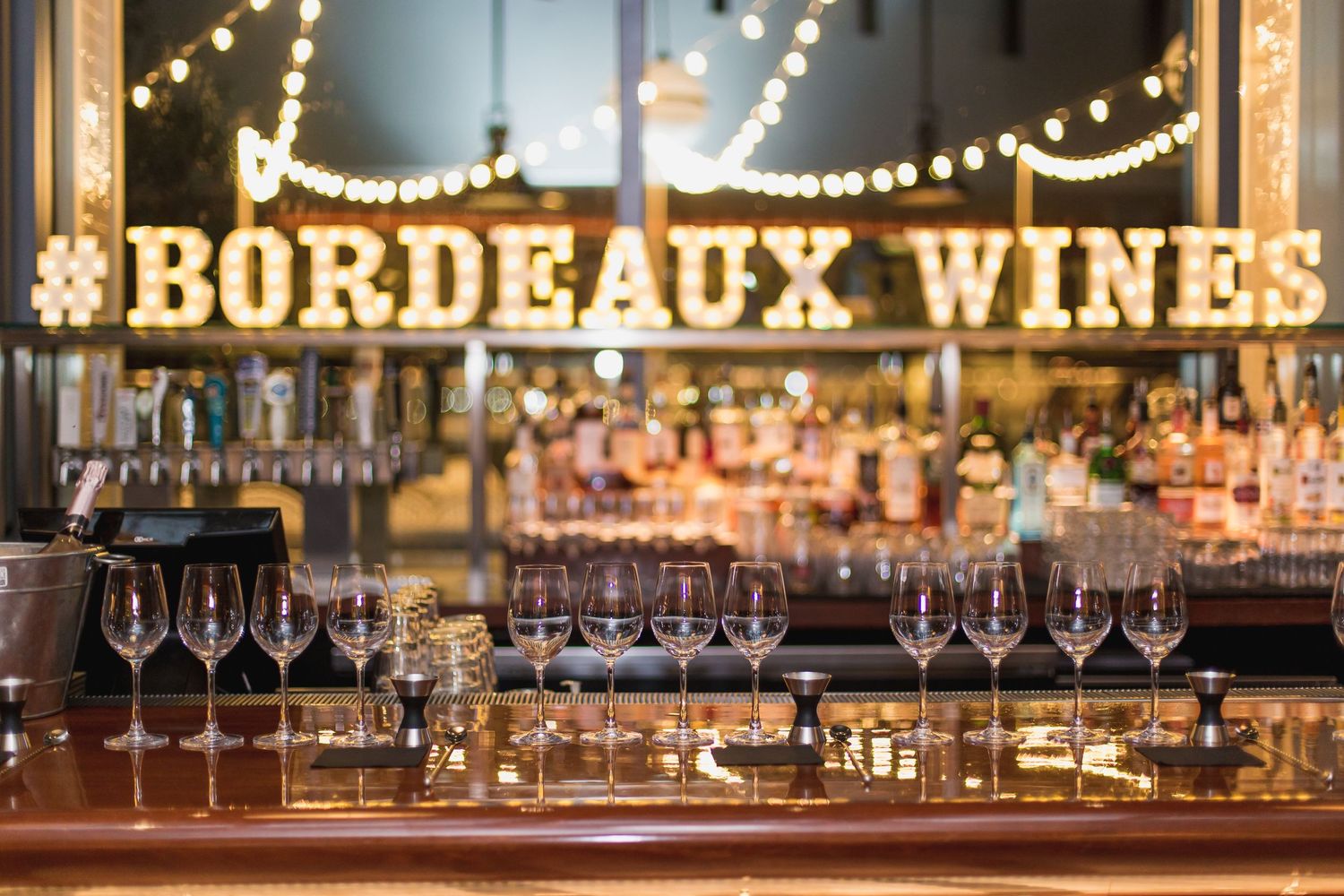Bar counter with wine glasses and illuminated Bordeaux Wines sign with string lights.