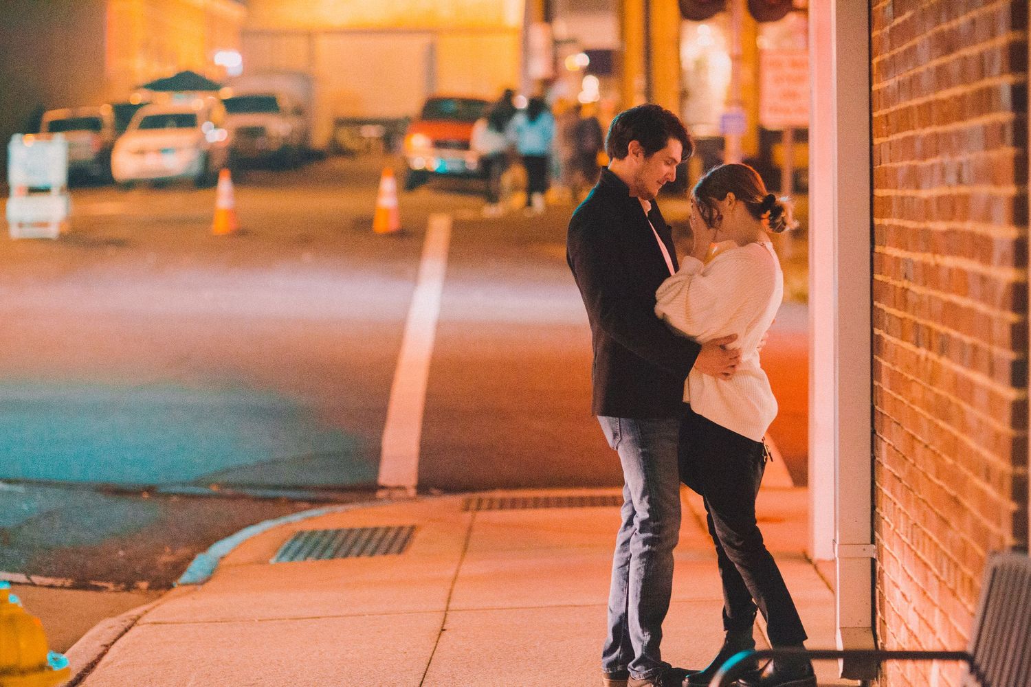A couple stands close together on an illuminated city street at night.
