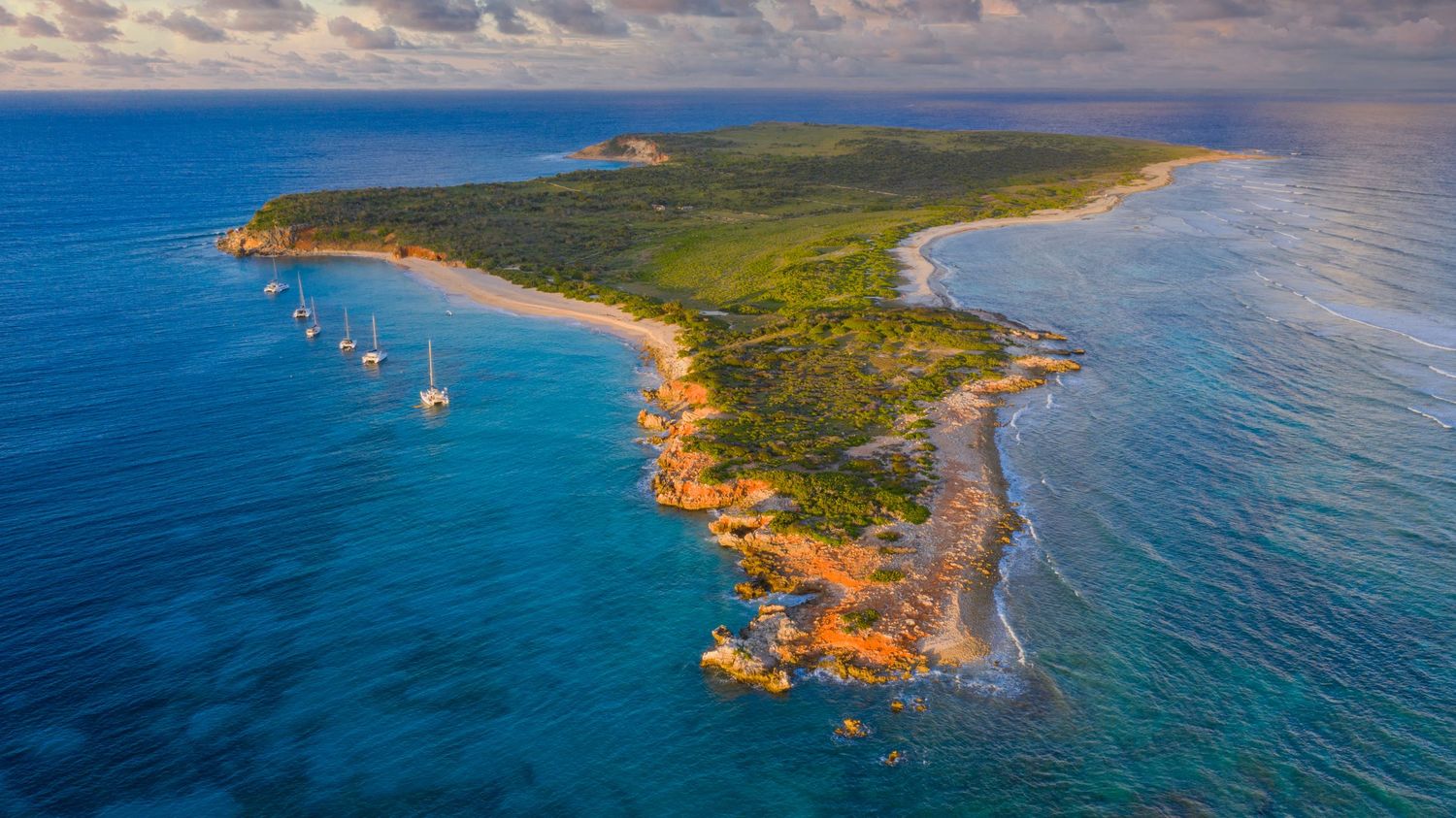 Aerial view of coastal peninsula with sailboats anchored along turquoise shoreline.