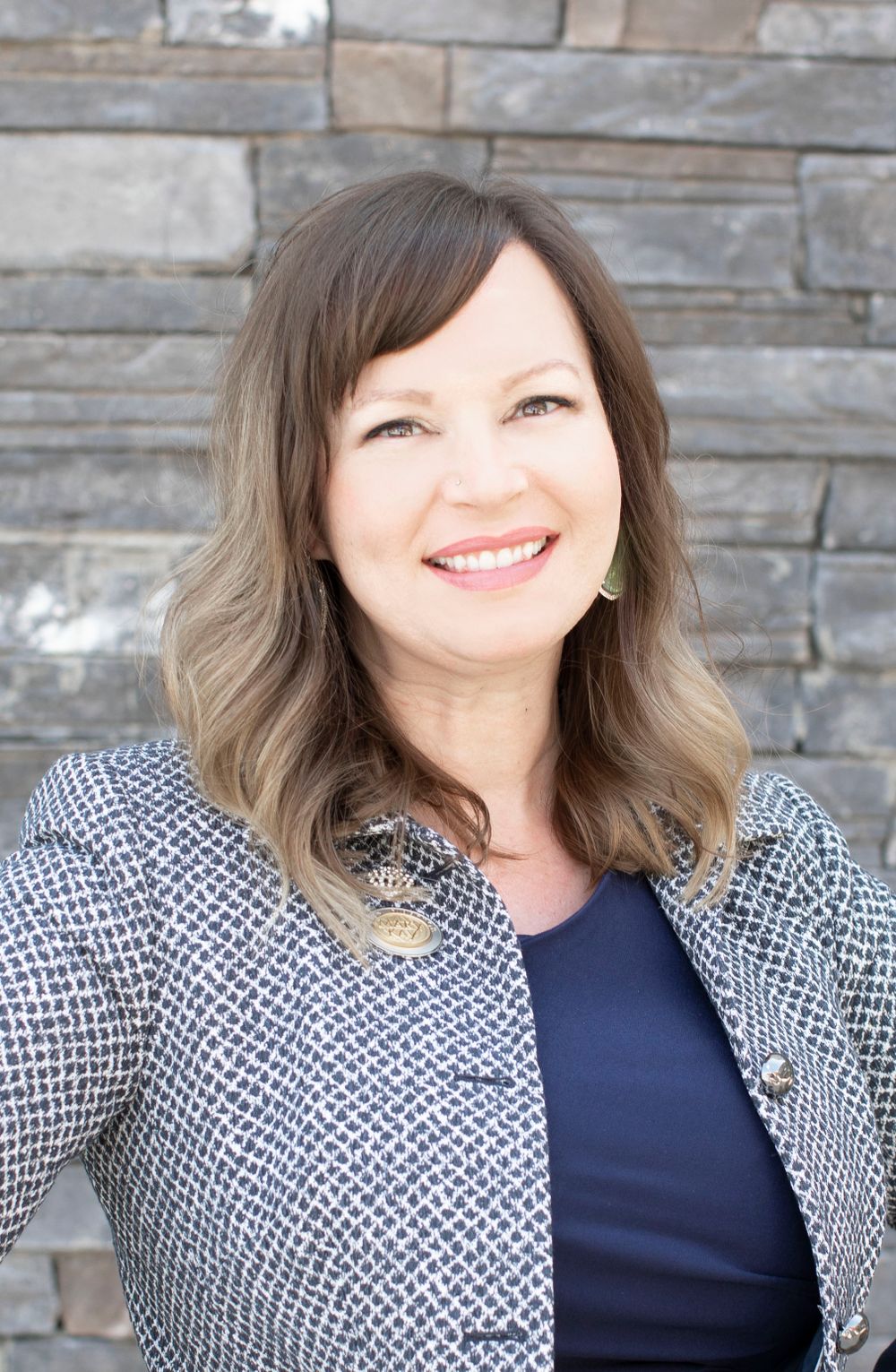 Professional headshot of woman in navy top and textured grey blazer against stone wall.