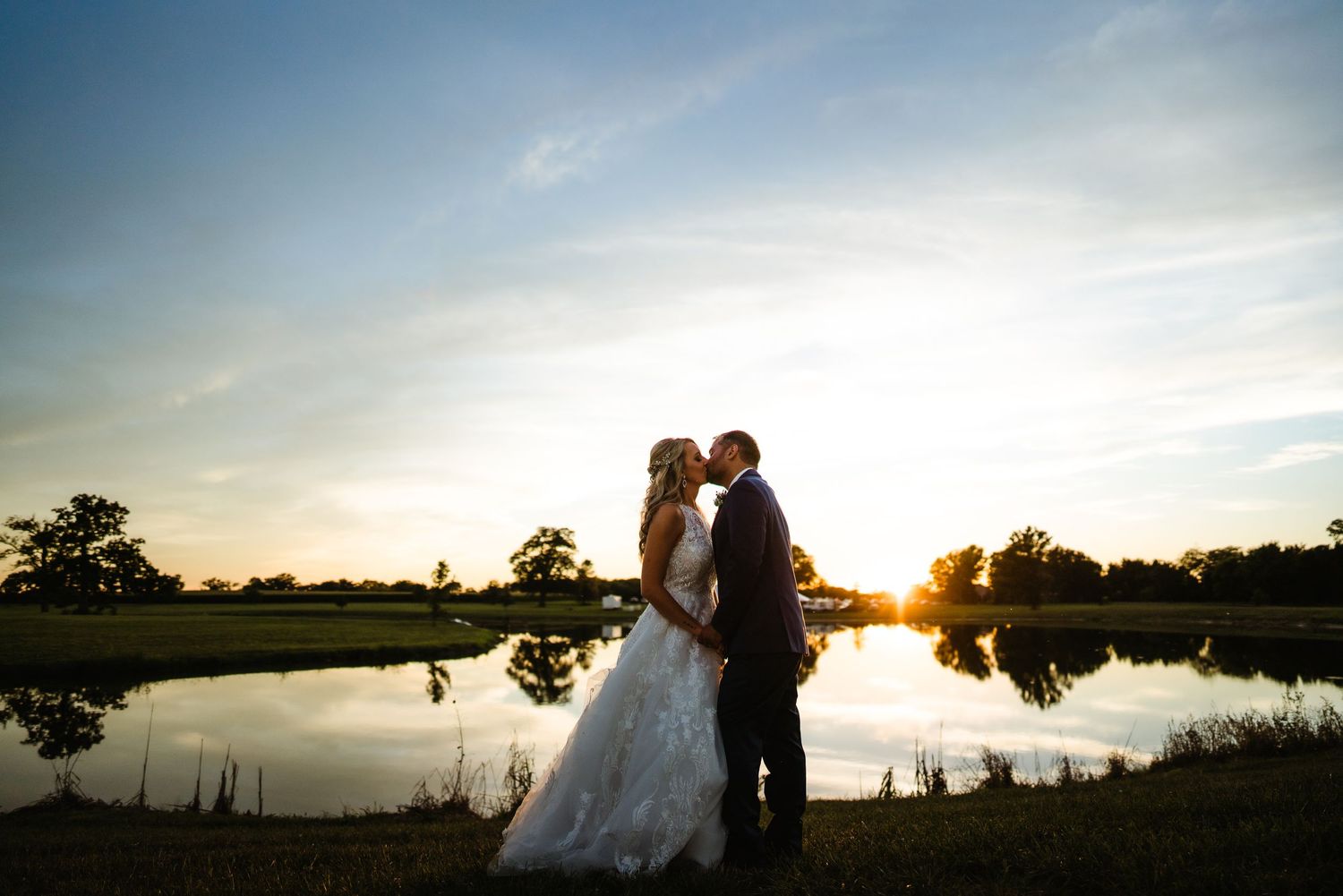 Romantic silhouette of a wedding couple embracing by a lake at sunset with golden sky reflections on the water.