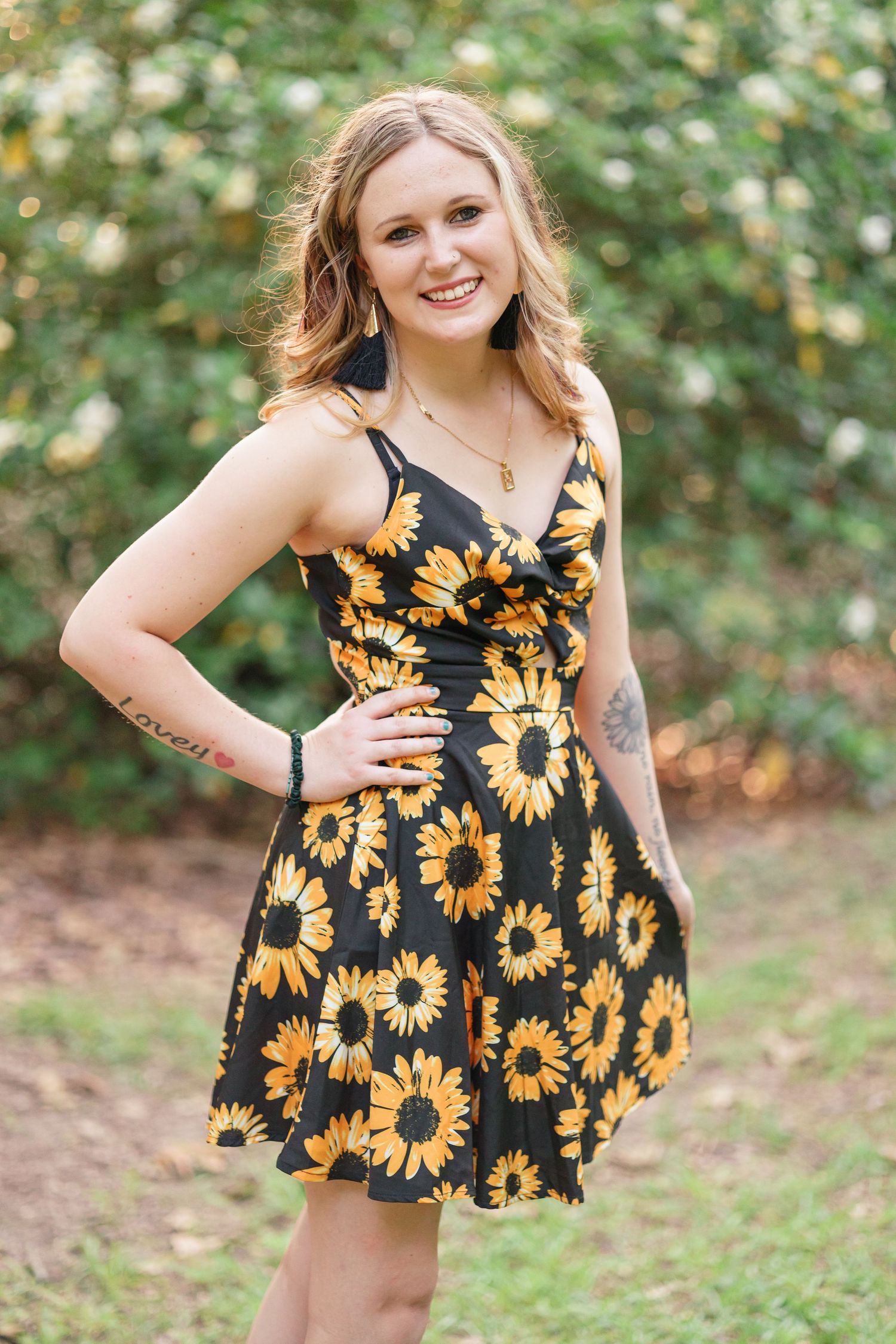 Woman in black and yellow sunflower dress poses outdoors in natural setting.