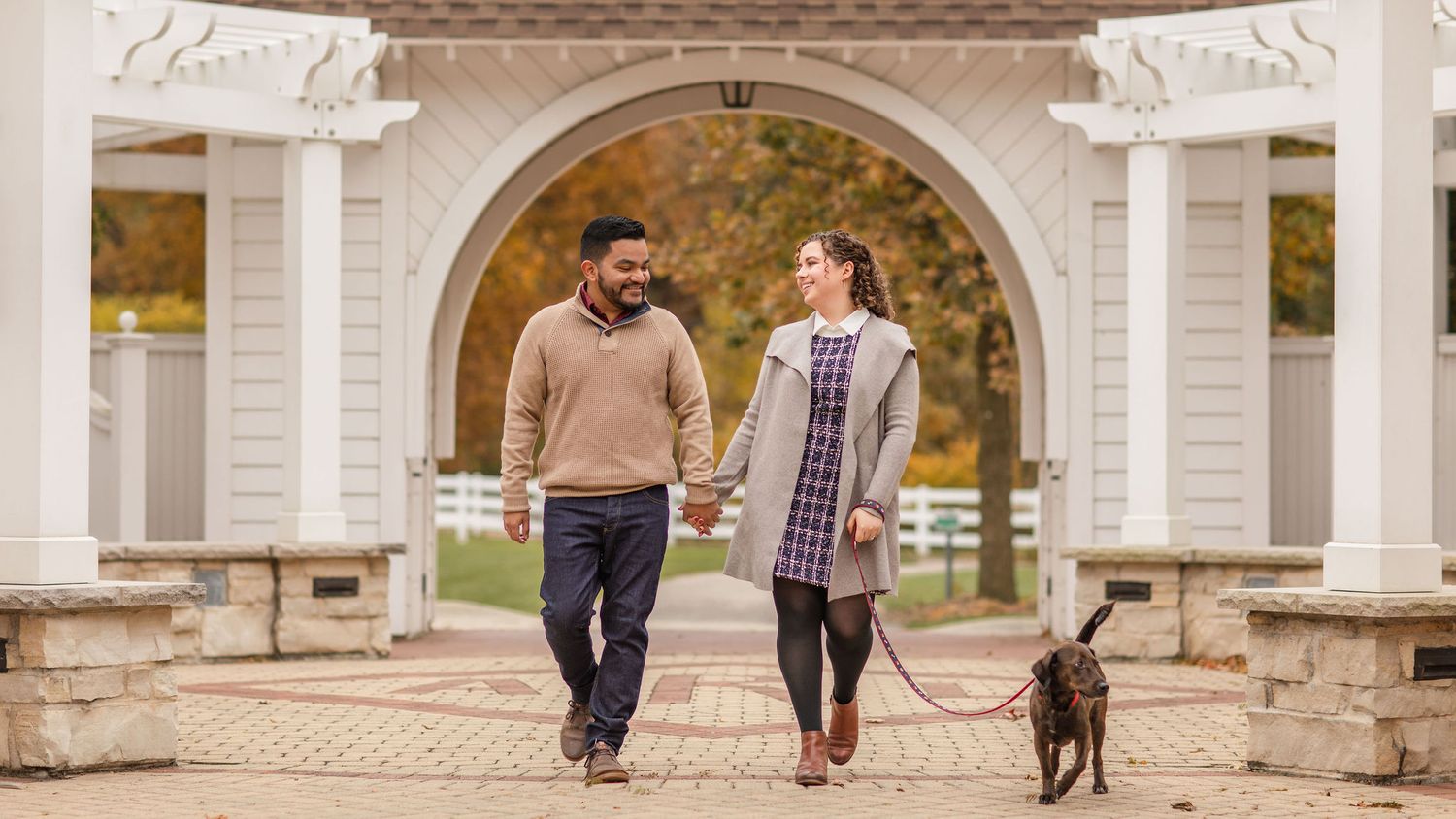 A couple walking through an ornate white archway with their dog on a leash.