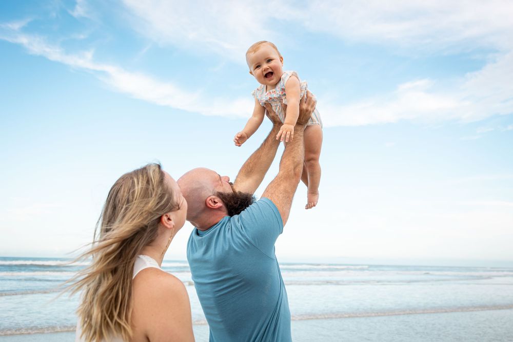 A joyful moment at the beach as a couple lifts their baby up against a bright blue sky.