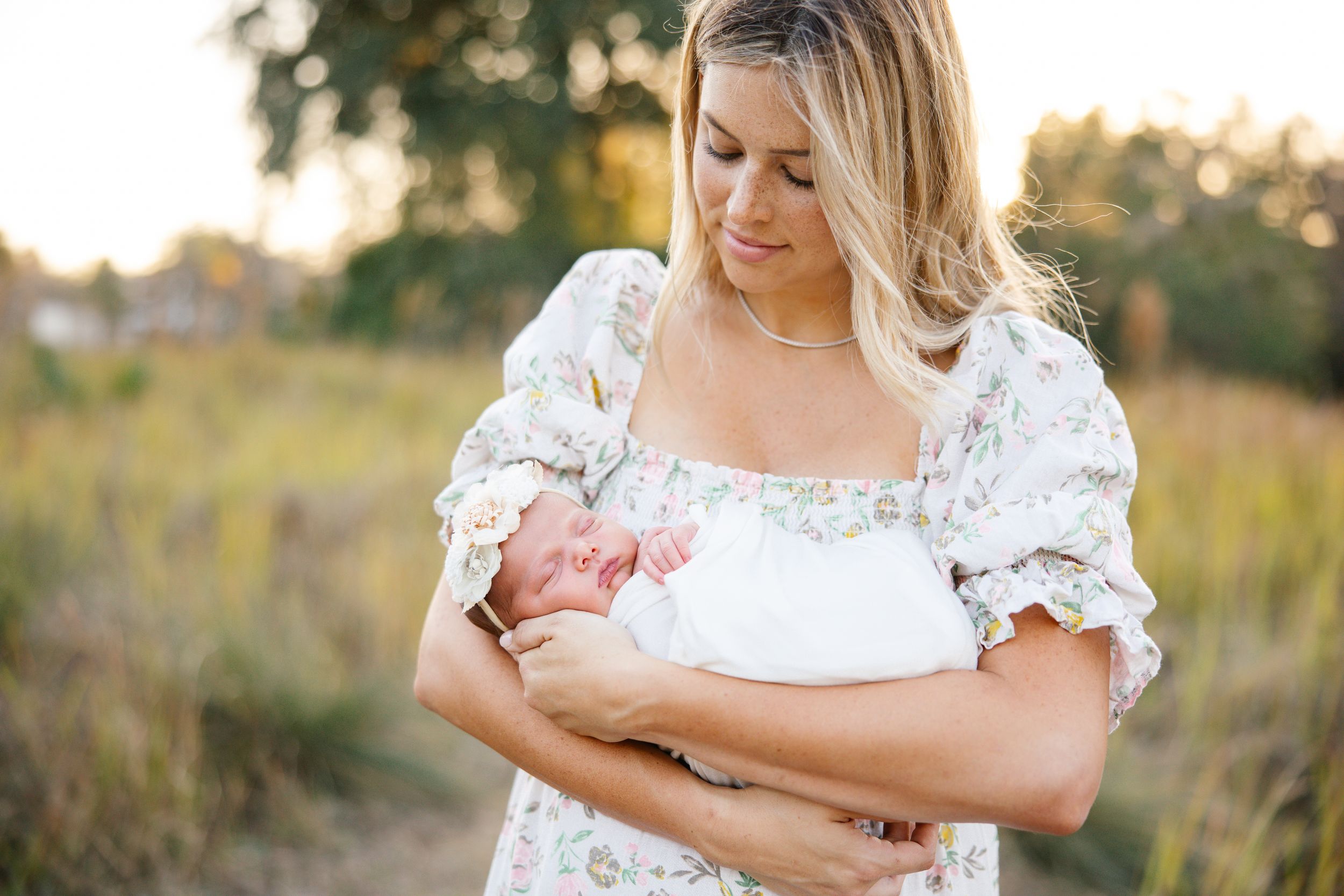 Woman in floral dress tenderly holding sleeping newborn baby outdoors in golden hour light.