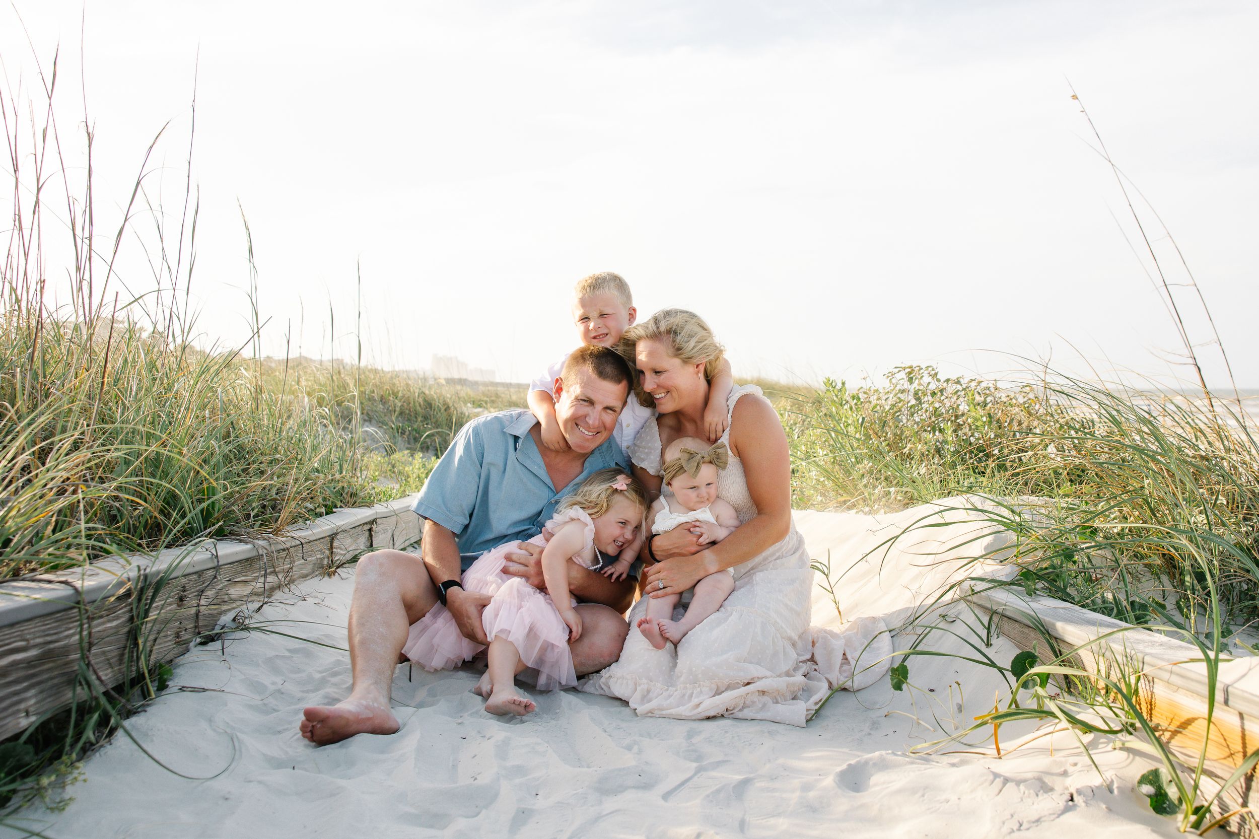 Family of five cuddling together on sandy beach dunes with tall grass, sharing a joyful moment outdoors.