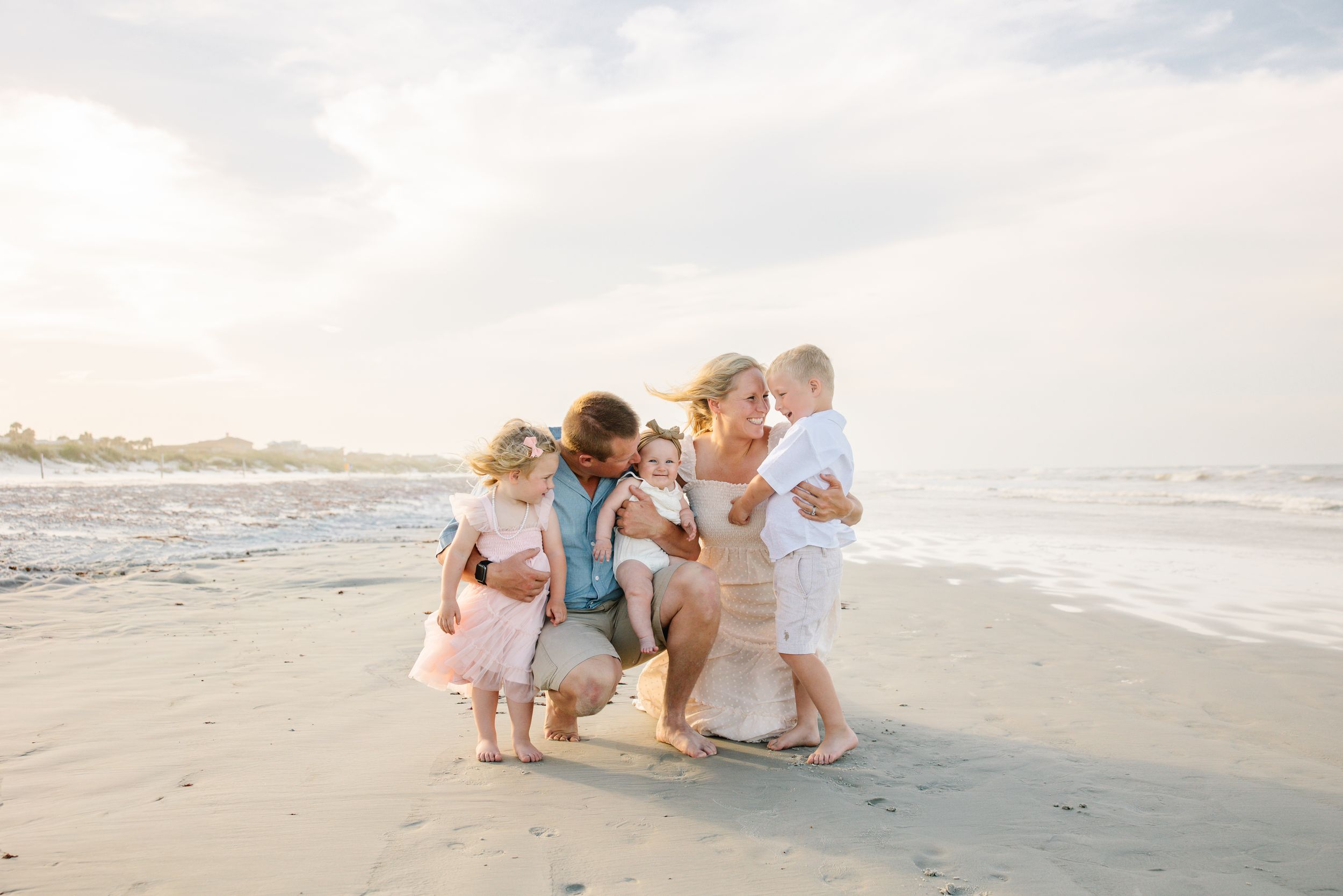 Family of five enjoying a sunny beach day, with parents and three young children playing together on the sandy shore.