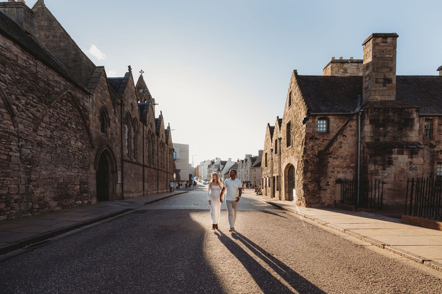 Silhouetted figures walk along a historic cobblestone street lined with old stone buildings at sunset.