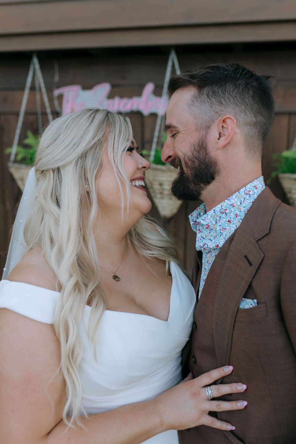 A joyful wedding couple laughs together in front of a pink neon sign.