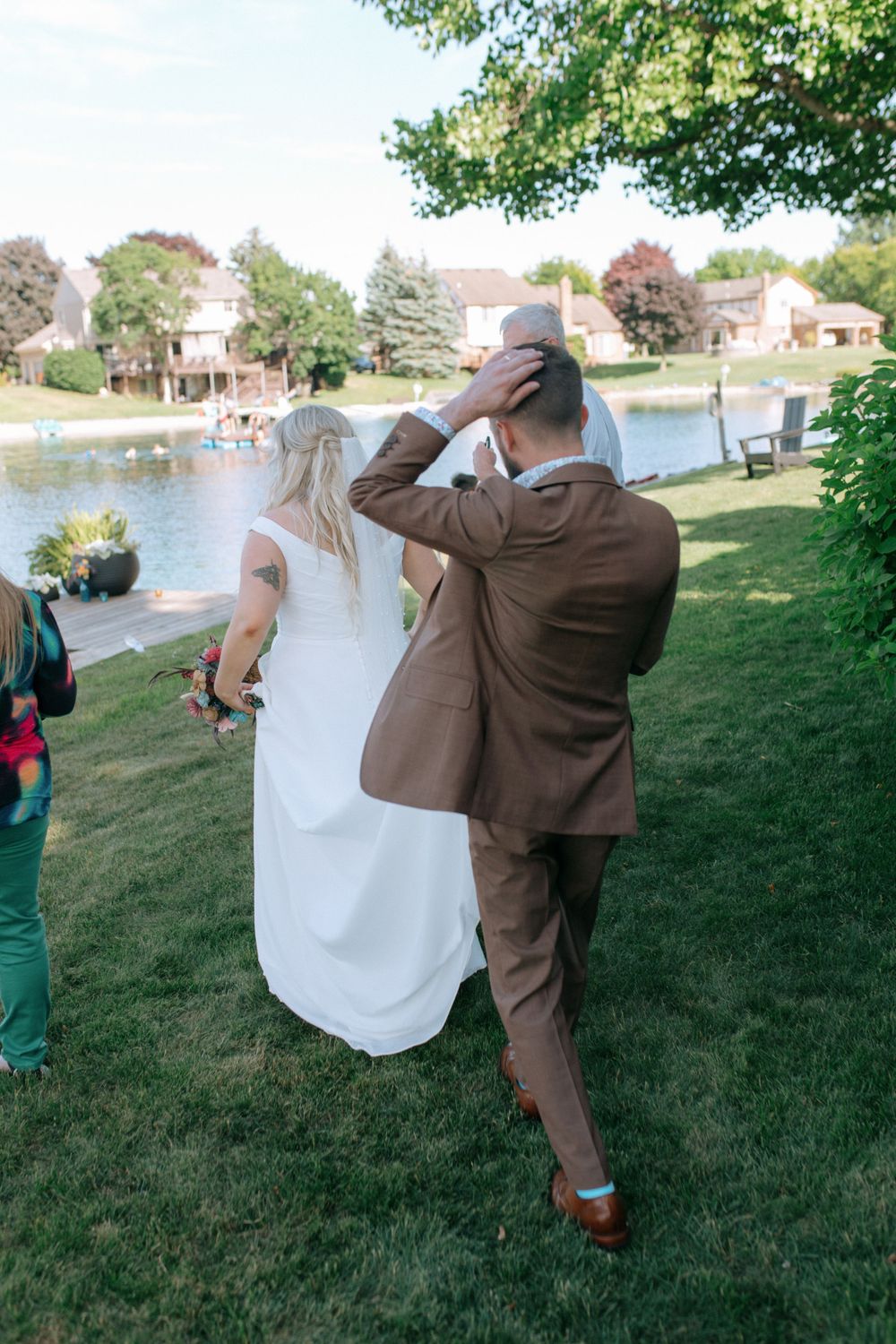 Wedding couple walking along grassy lakeside path with the groom in brown suit.