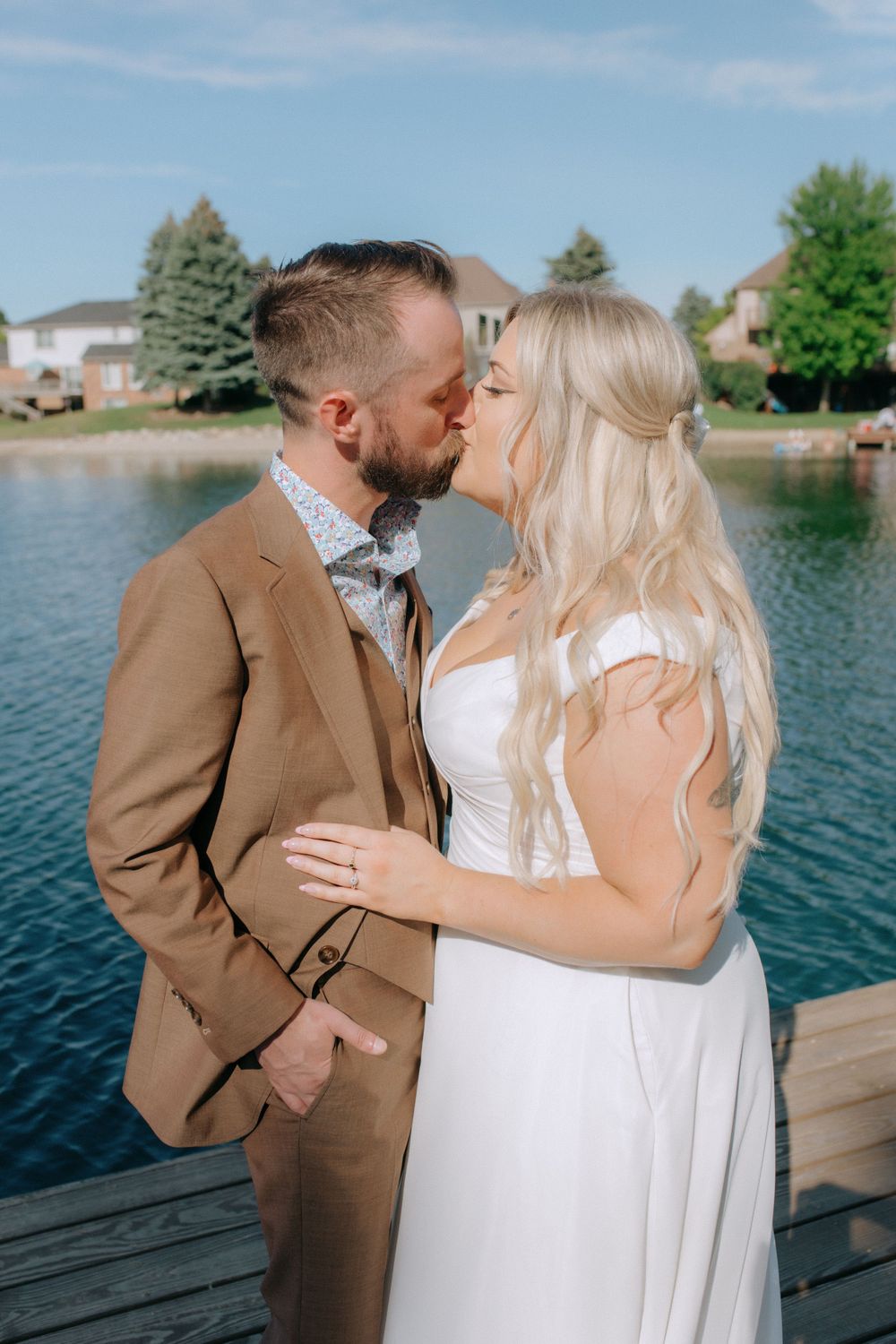 Couple shares romantic kiss on wooden dock with lake and blue sky backdrop.