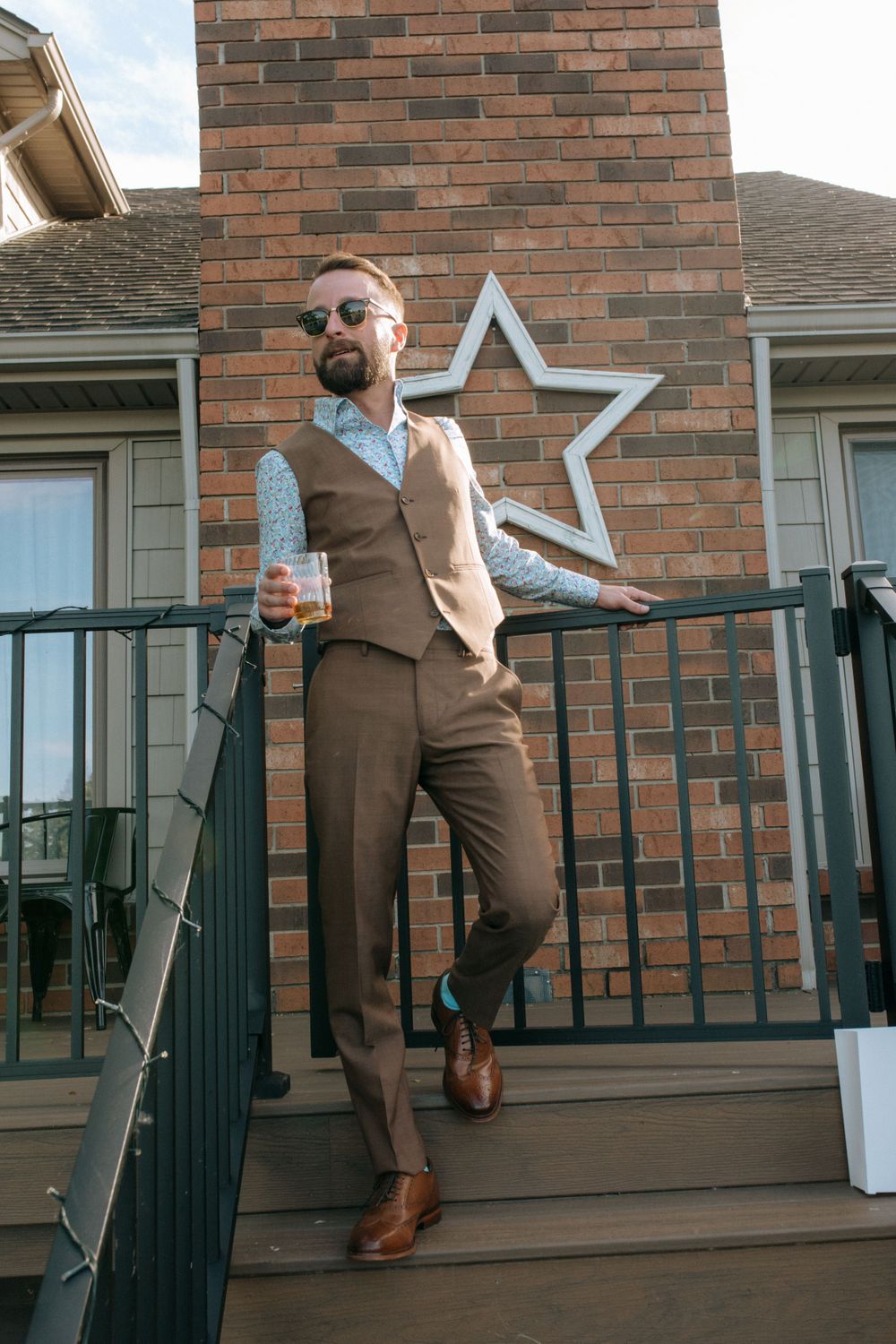 Stylishly dressed person in brown suit and vest poses casually on porch with decorative star wall accent.
