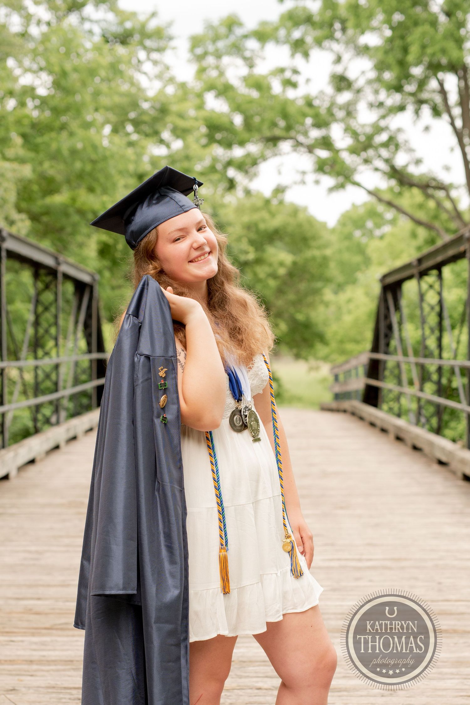 A graduate in white dress and cap poses with gown draped over arm on a wooden bridge.