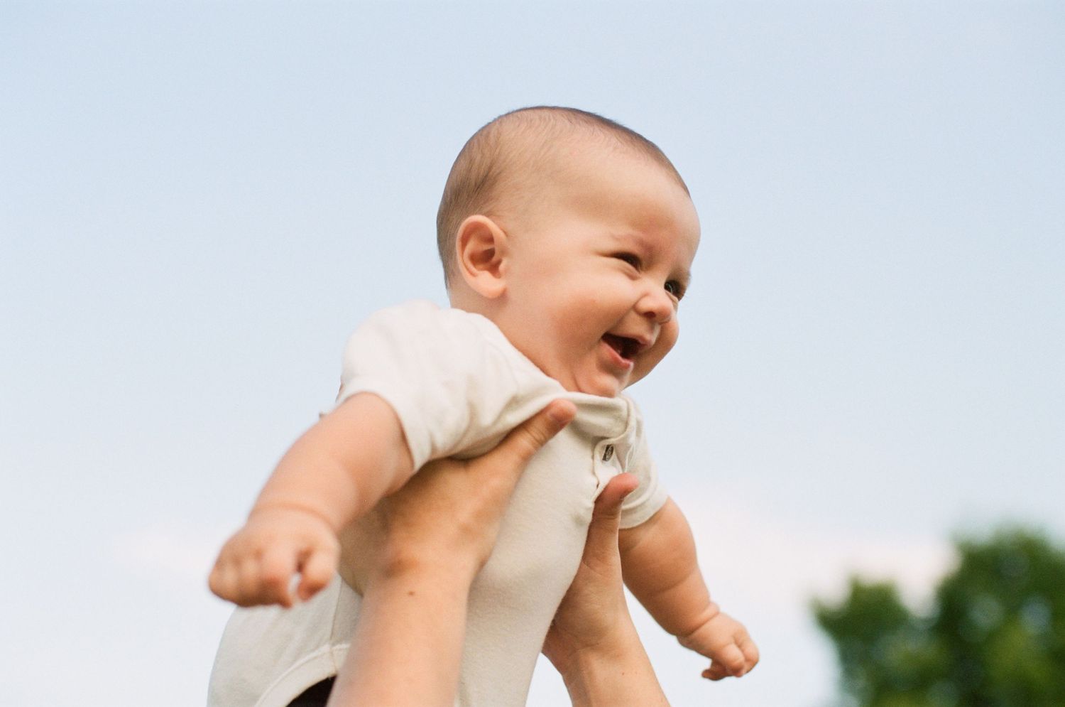 Baby in white onesie laughing while being lifted up against blue sky.
