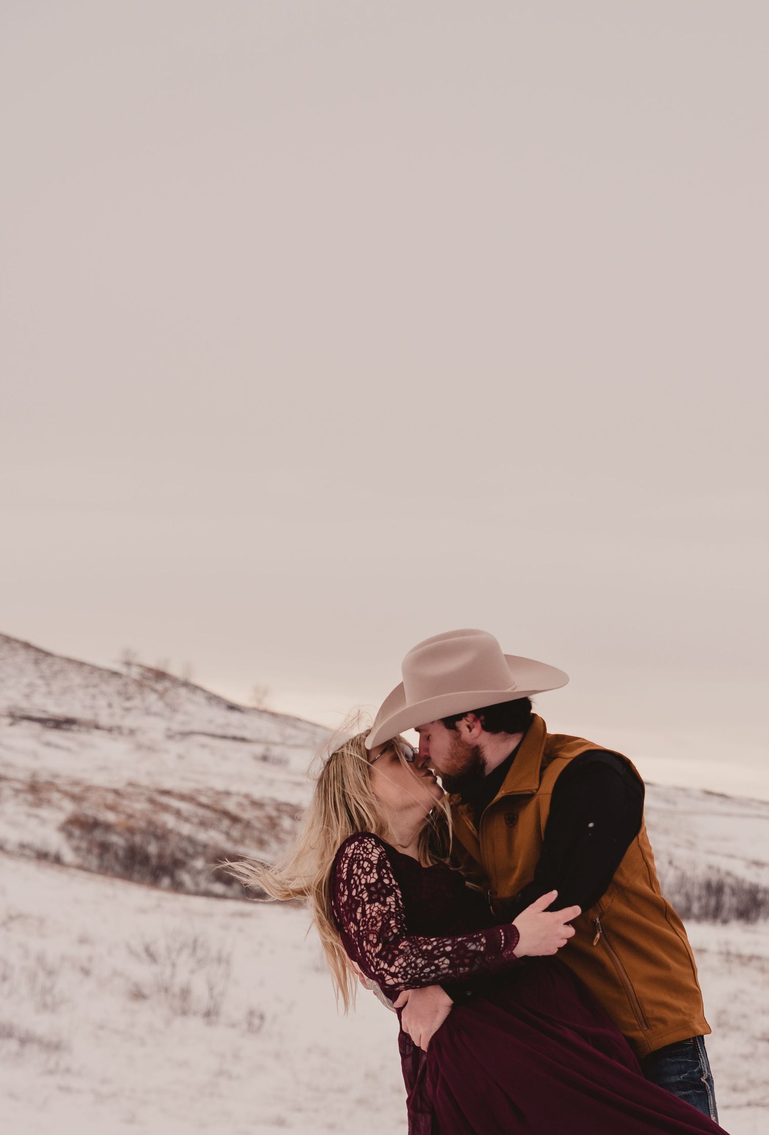 Romantic winter portrait session with couple in burgundy dress and brown jacket against snowy background.