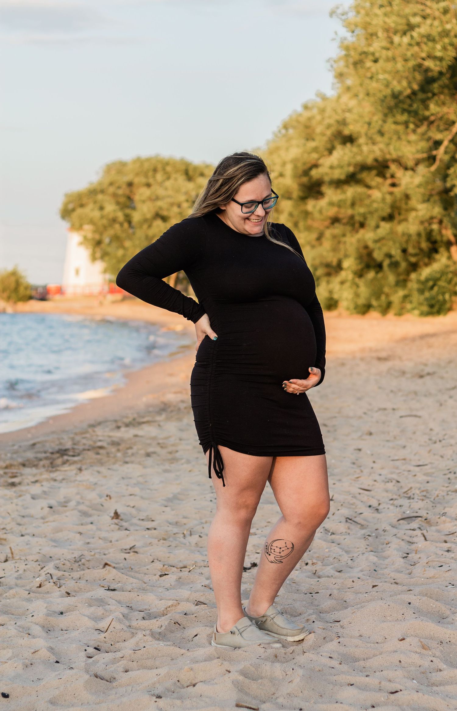 Pregnant woman in black dress poses confidently on beach during sunset photoshoot.