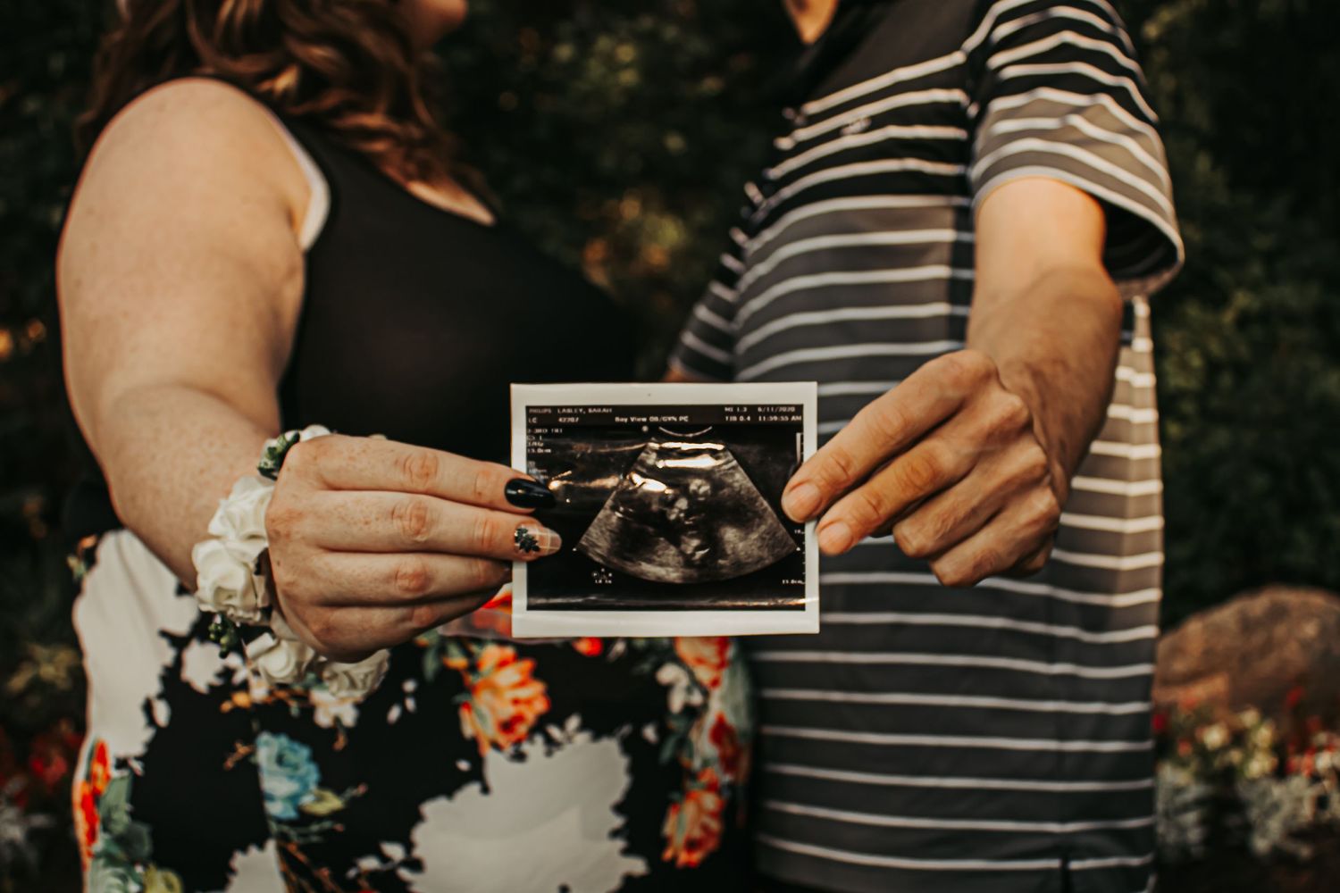 A couple holding up an ultrasound photo in front of garden flowers.