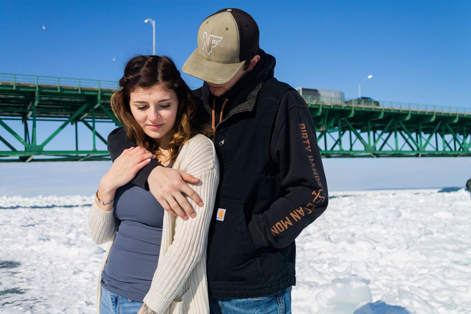 Intimate winter portrait of a couple standing in the snow near a large green bridge.