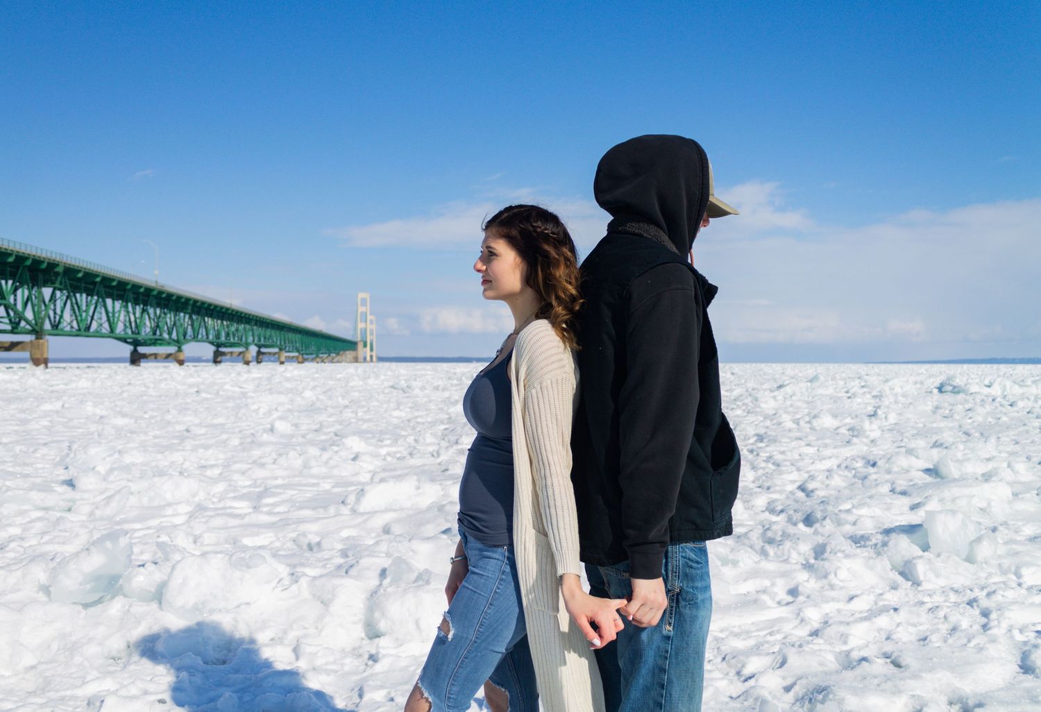 Couple holds hands while walking through deep snow with a large green bridge in the background.