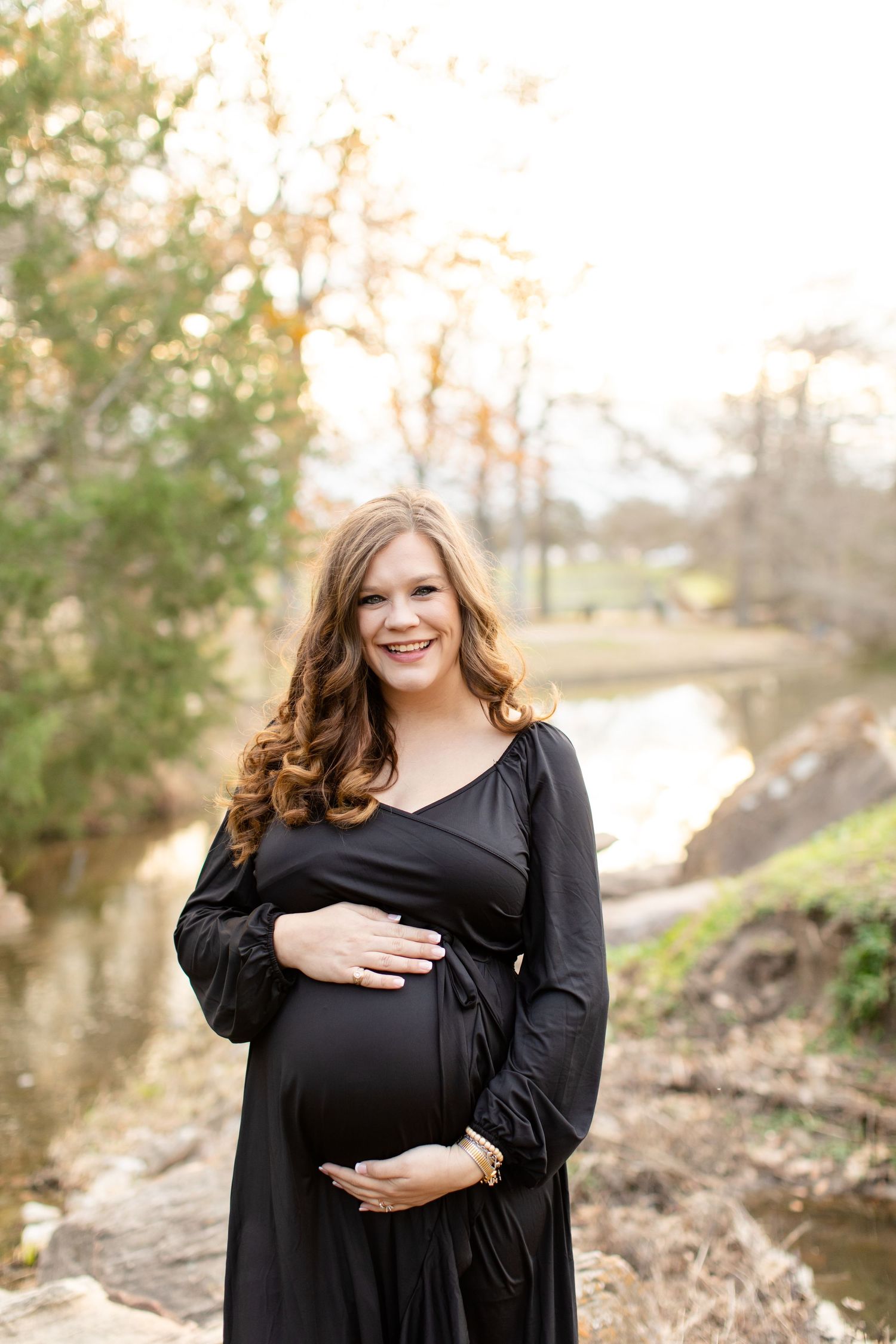 Expectant mother in black dress smiling by creek during autumn photoshoot.
