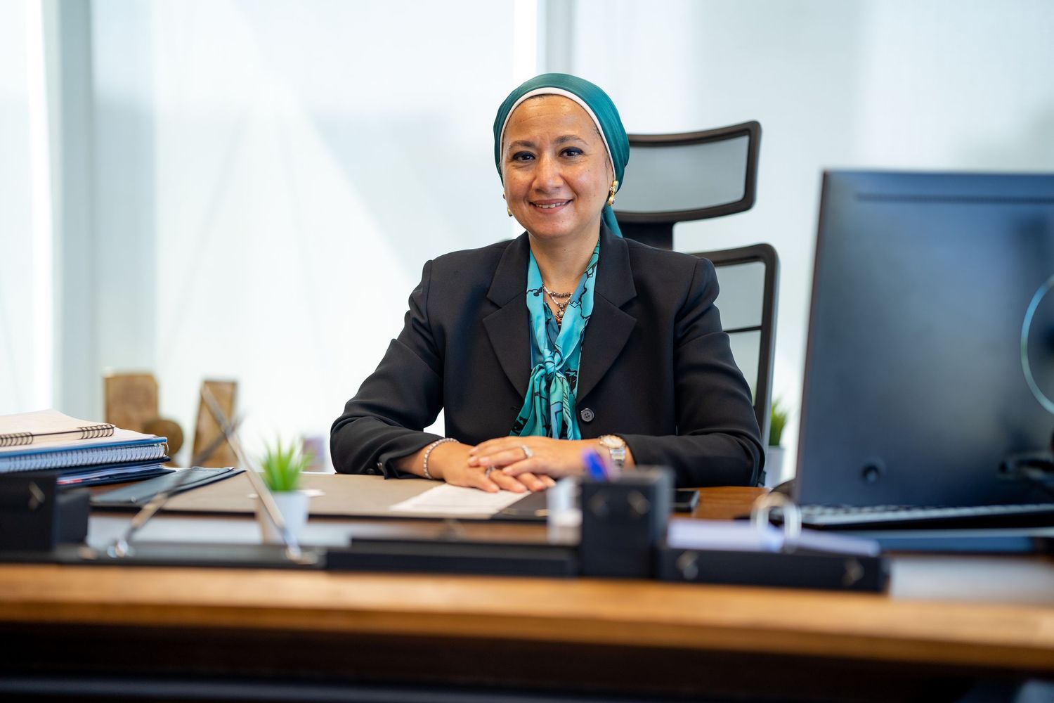Professional at desk wearing business attire and headscarf in bright office setting.