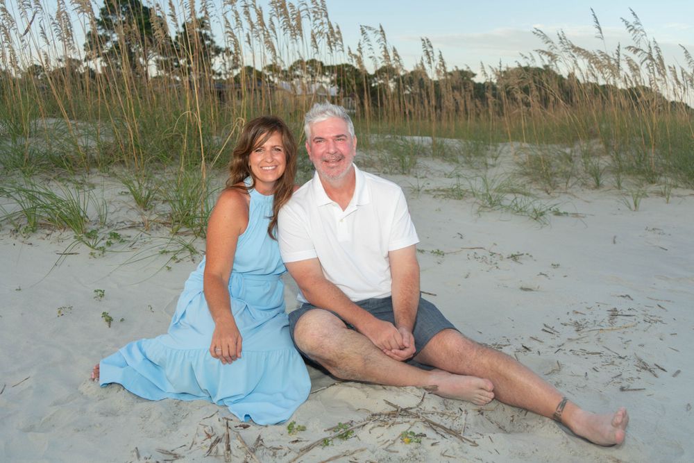 Smiling couple sitting together on sandy beach with sea oats in background at sunset.
