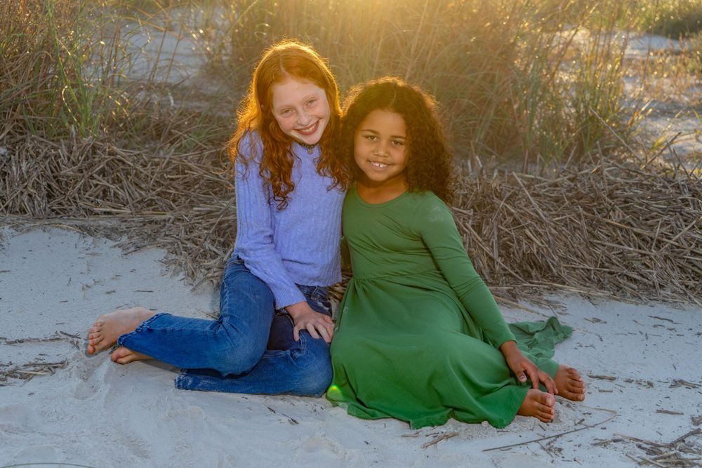 Two girls sitting on sandy beach dunes smiling in golden hour light.