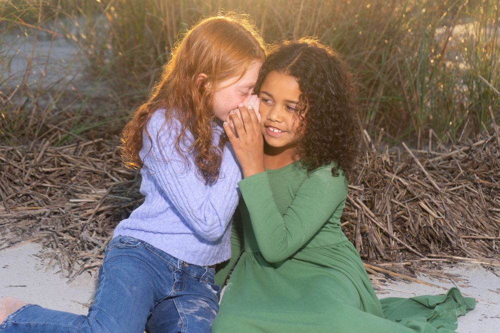 Two girls whispering and sharing secrets while sitting on sandy beach.