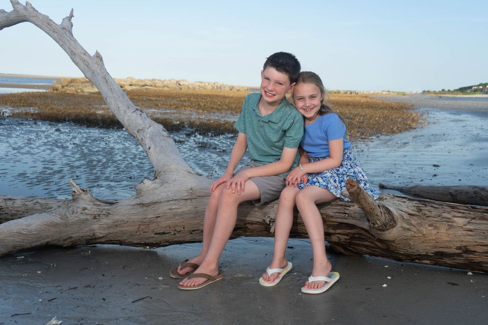 Two children sitting together on driftwood log at beach shoreline, smiling at camera.