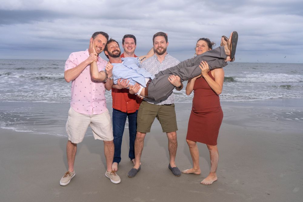 Group of five friends playfully carrying one person on beach with ocean waves behind them.