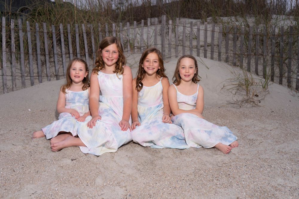 Four young girls in matching white dresses posing on beach sand.
