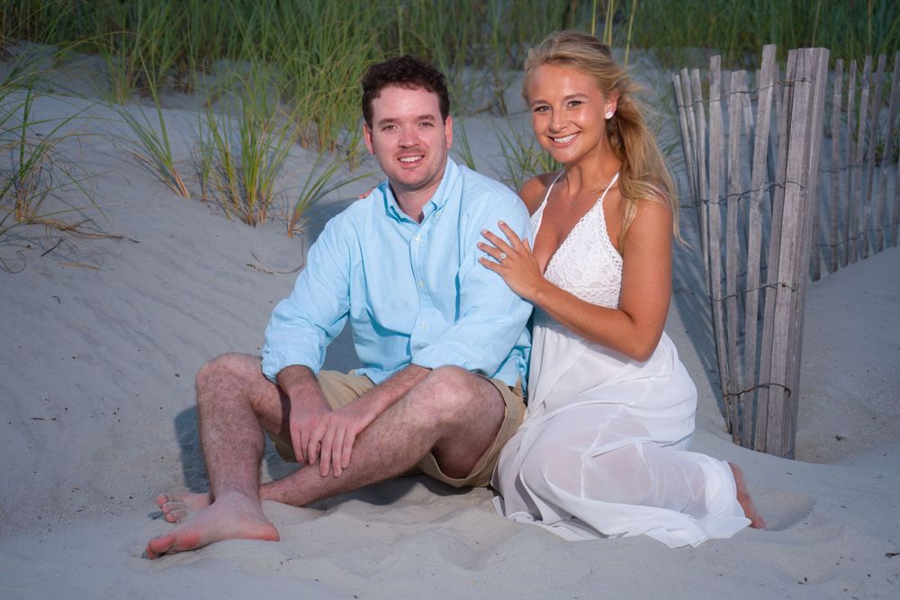 Engaged couple sitting on beach sand dunes at sunset, woman showing off engagement ring.