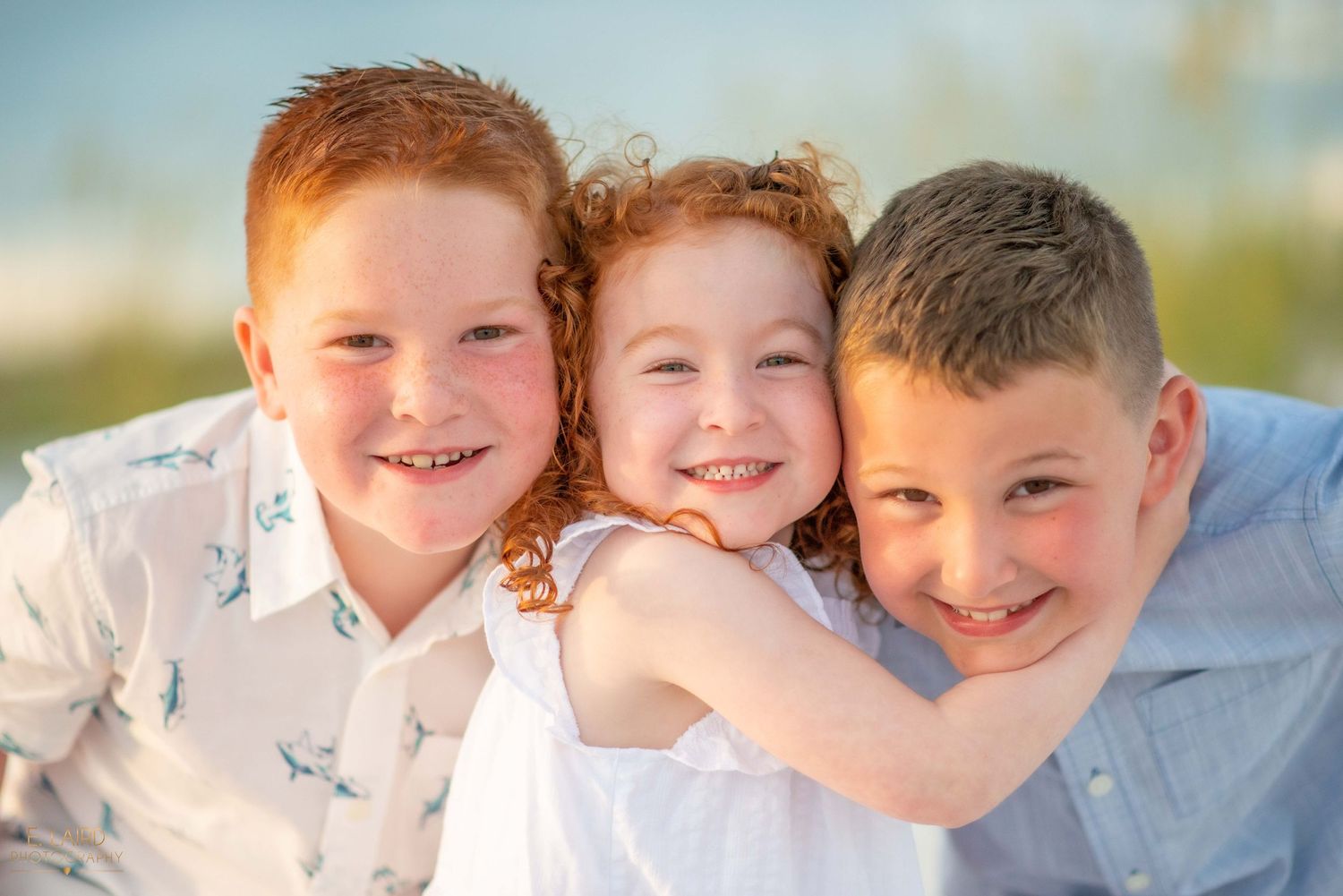 Three children share joyful embrace against blue sky backdrop.