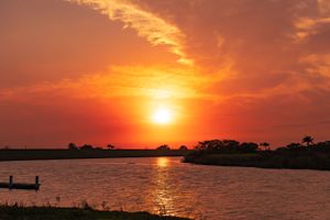 Vivid orange sky at sunset over a tranquil waterway with distant trees on the horizon.