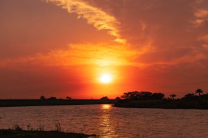 Glowing orange sun descends over a serene waterway with silhouetted landscape.