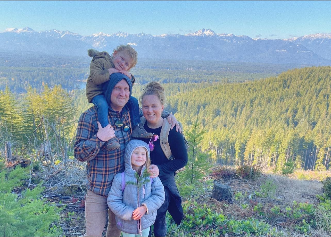 A group stands together on a mountain overlooking a vast evergreen forest with the snow-capped peak Olympic Mountains in the distance.
