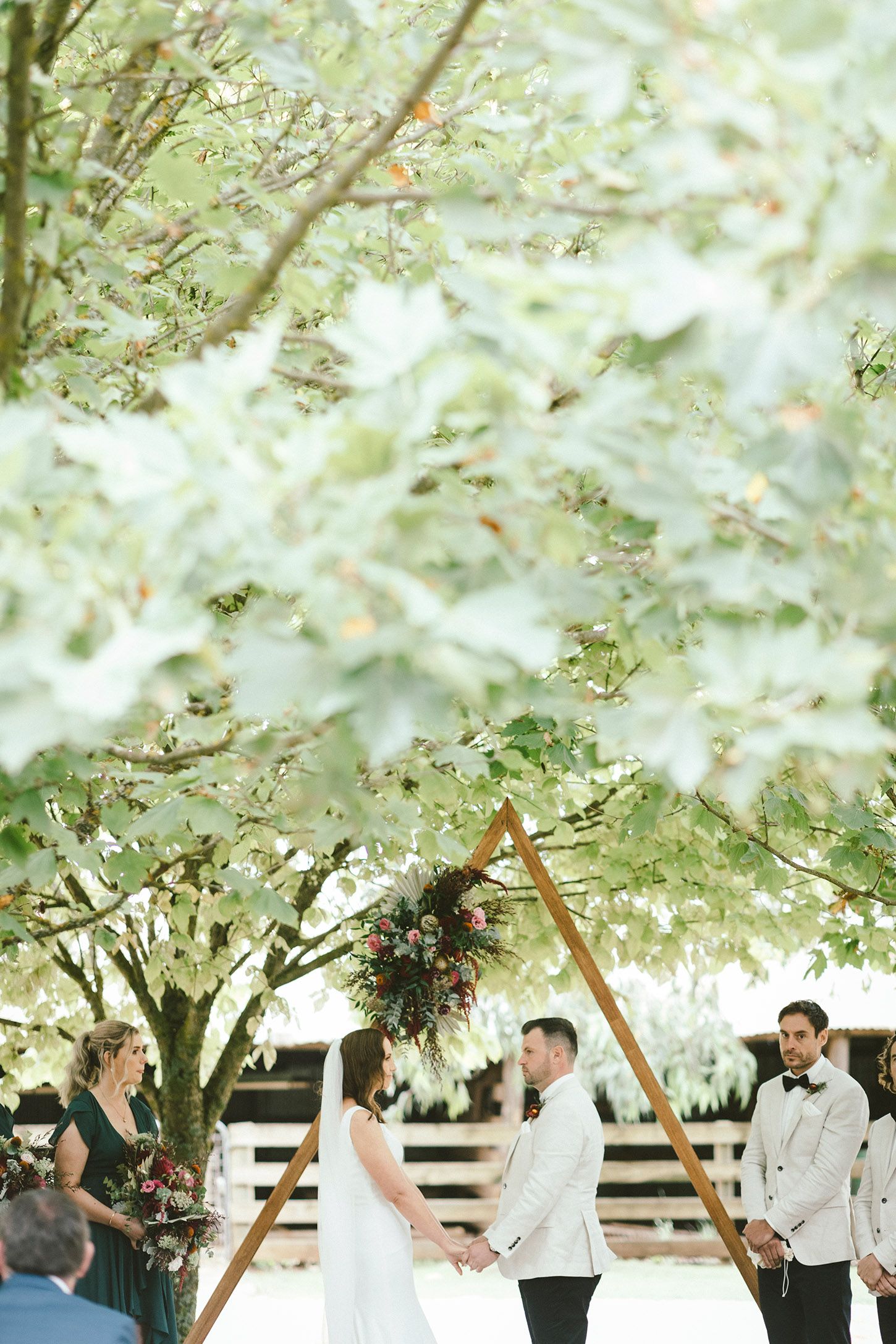 Wedding ceremony under blooming trees with wooden triangle arch and white floral decorations.