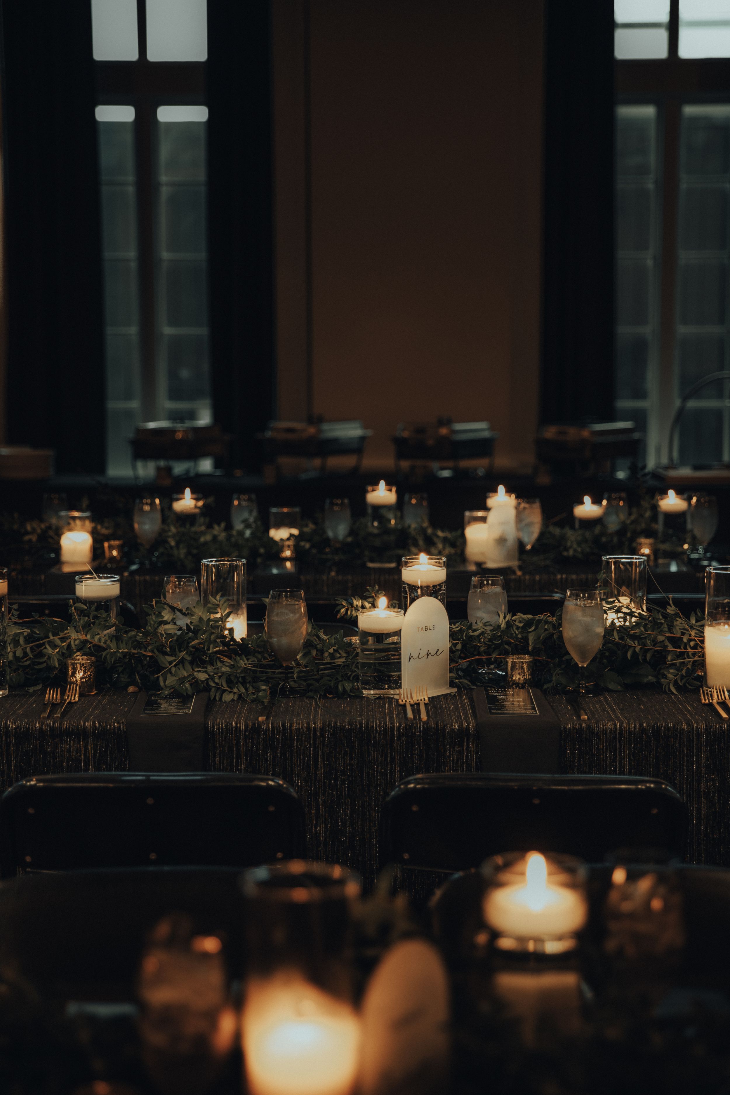 Atmospheric wedding reception table decorated with glowing candles and greenery in dark lighting.