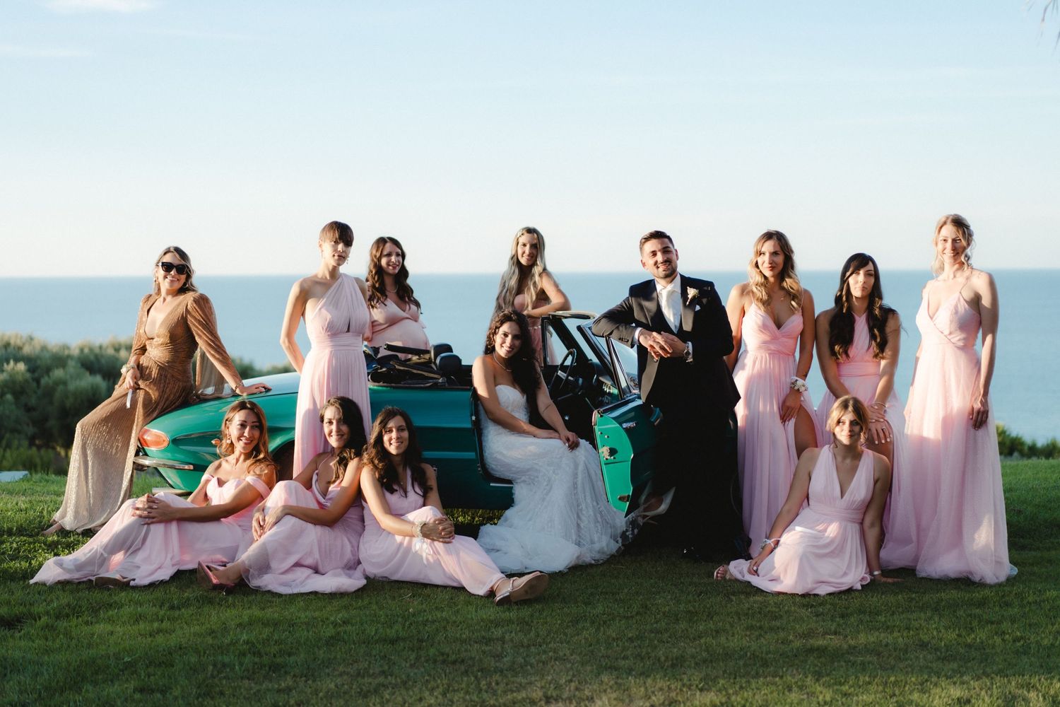 A wedding party in pink dresses poses with a vintage car against an ocean backdrop.