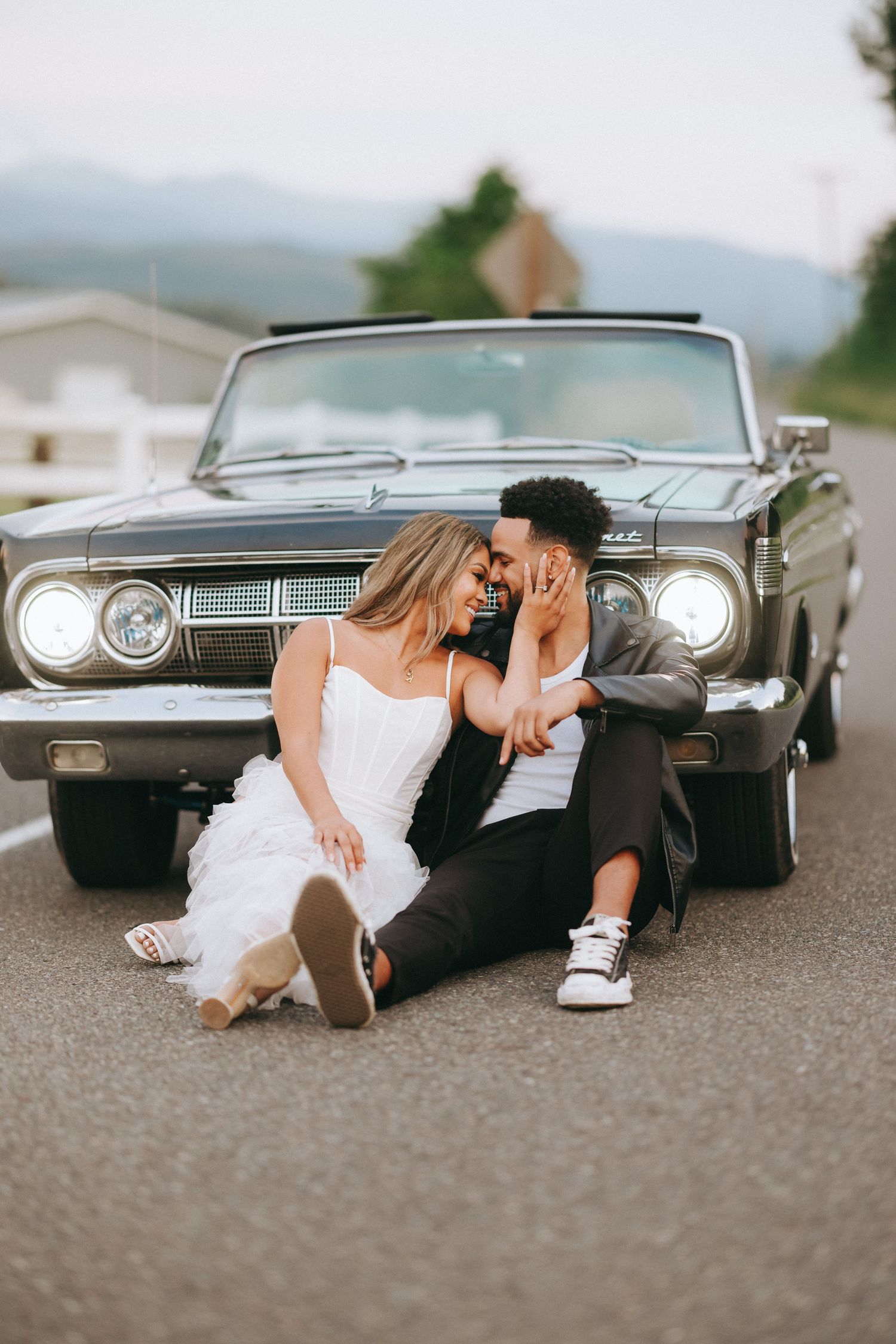 A romantic couple sits on the ground in front of a vintage black convertible car with headlights on.