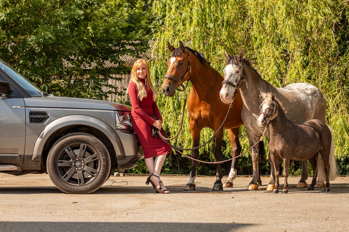 Three horses stand next to a modern SUV on a sunny day.