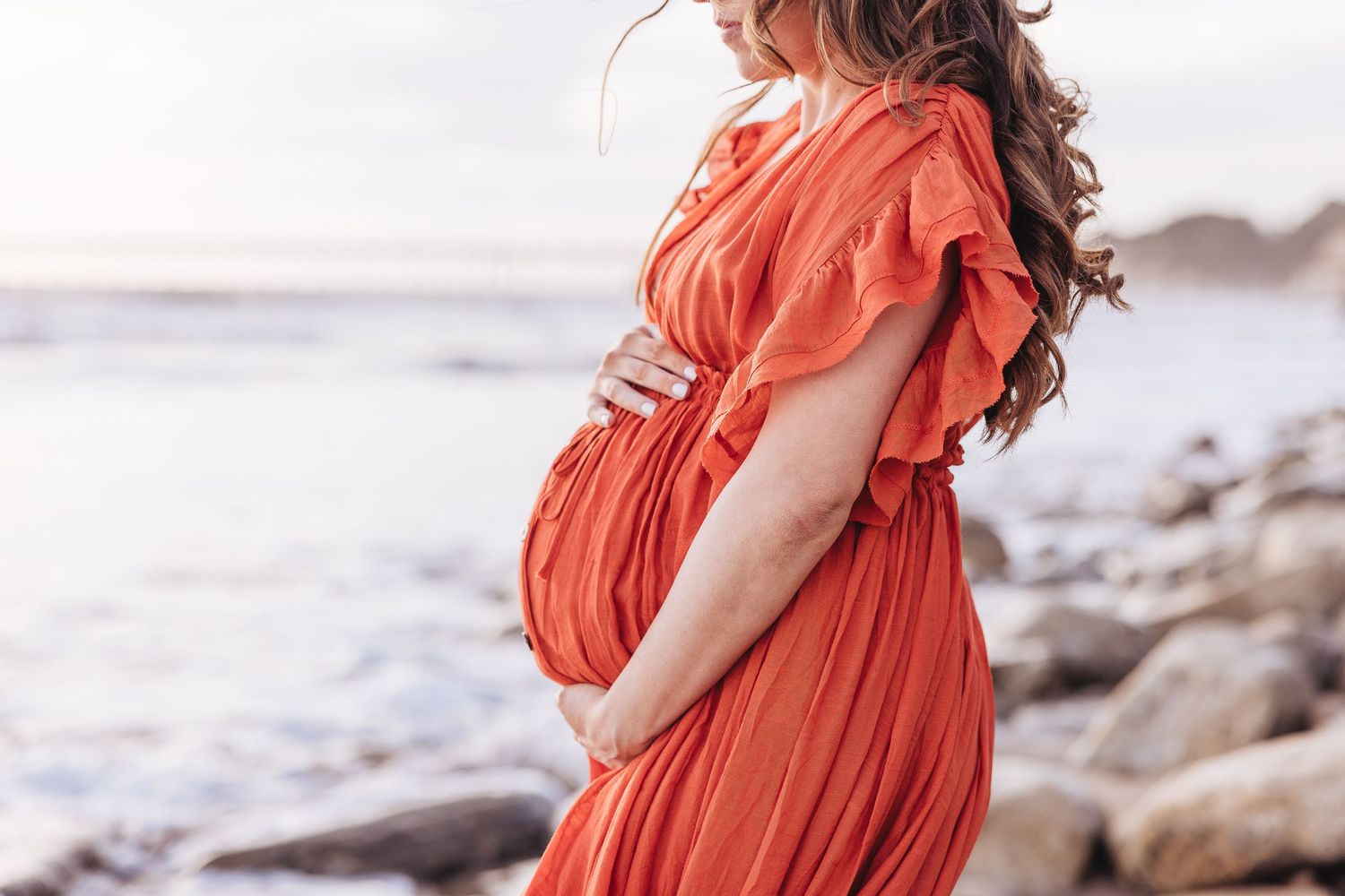 Flowing coral maternity dress highlighted against rocky coastal background.