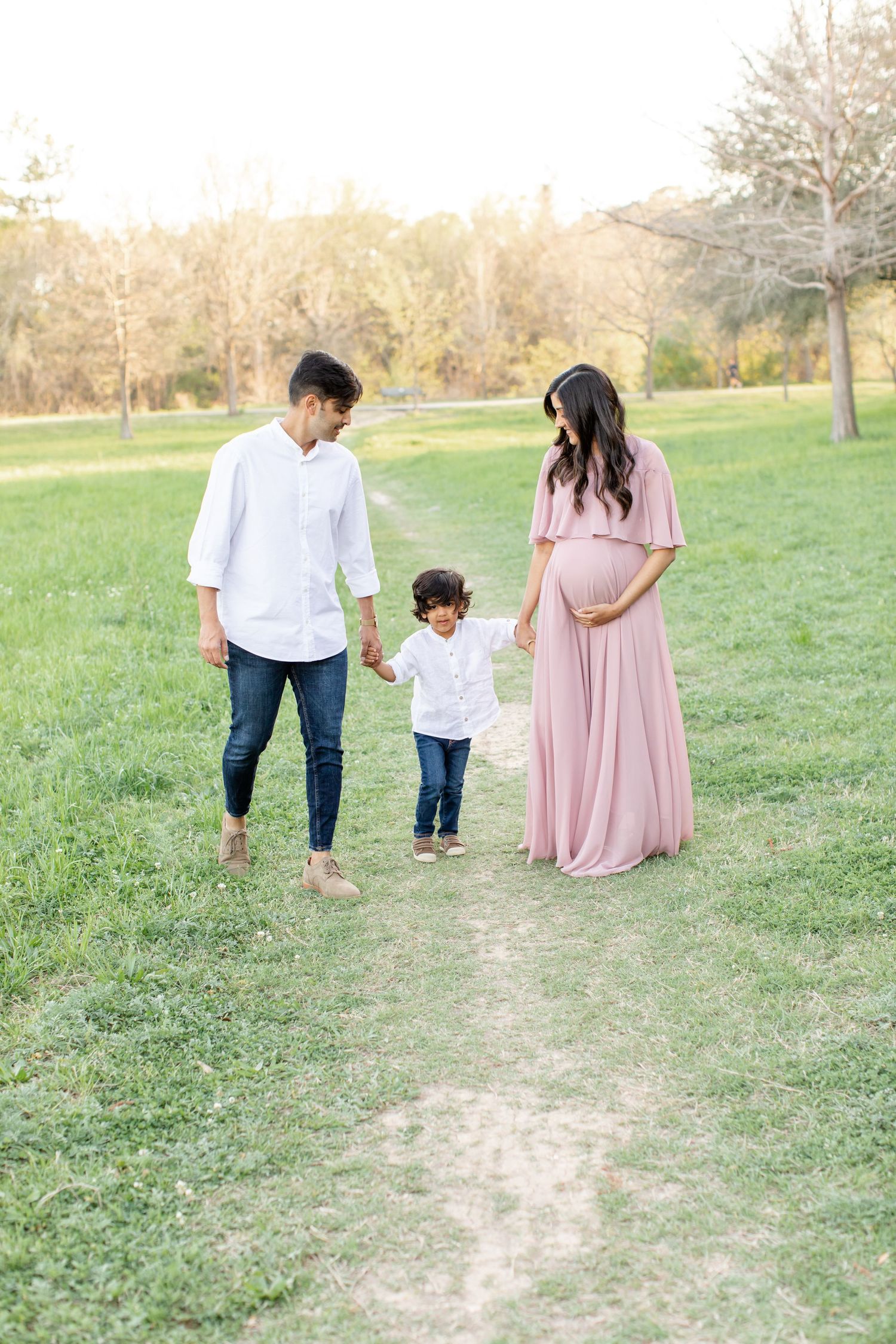 A family walks together on a path through a sunny park, with the woman wearing a flowing pink dress.