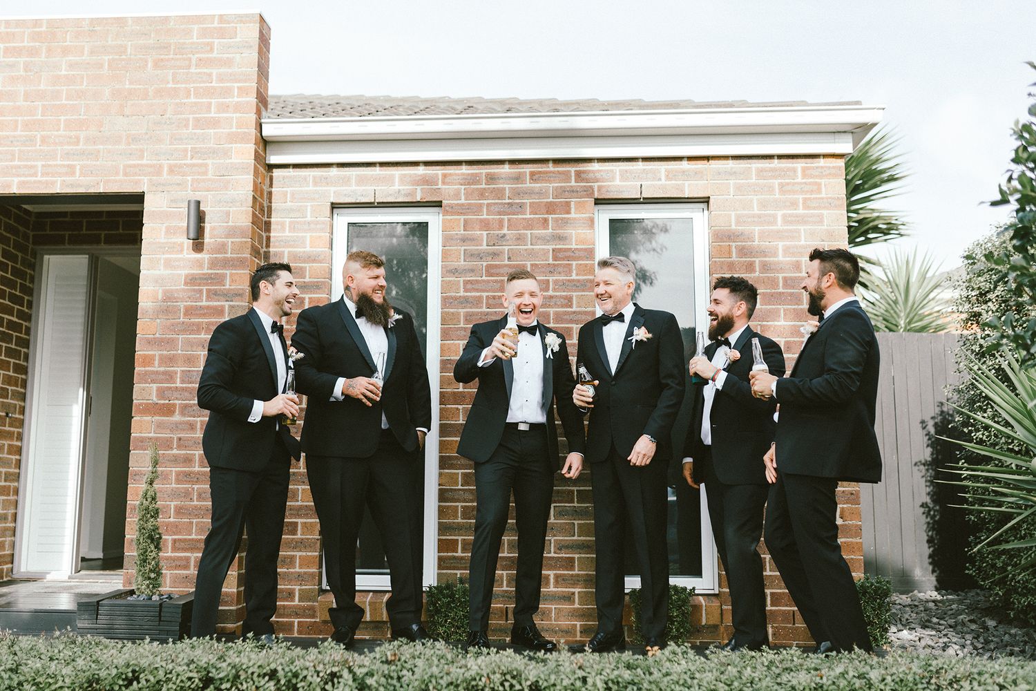 Groomsmen share laughs while posing together outside a brick building.